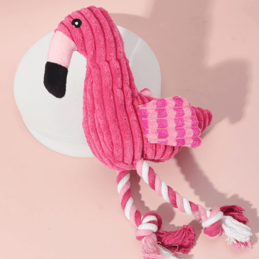 A pink flamingo dog toy with a squeaker inside. The toy is made of soft, plush material and is sitting on a white table. The flamingo has a long neck and legs, and it is pink in color. 