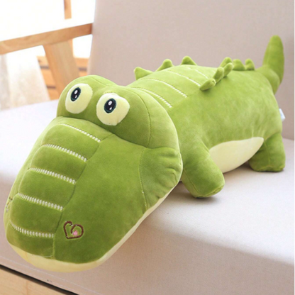 A stuffed crocodile toy is laying on a couch. The crocodile is green and has a long tail. It is made of soft plush material and has a closed mouth.