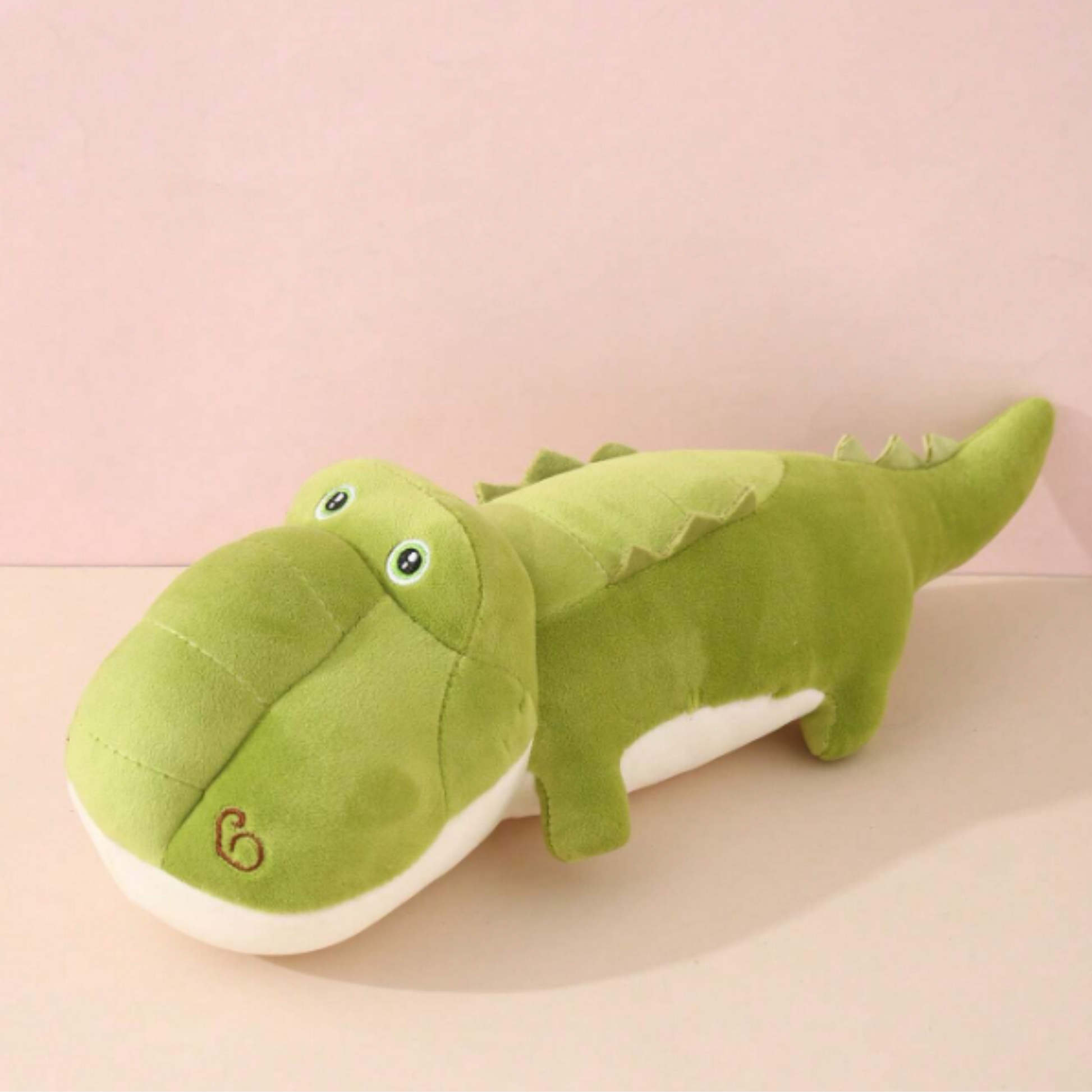 A stuffed crocodile toy sitting on a table. The crocodile is green and has a long tail. It is made of soft plush material and has a closed mouth.