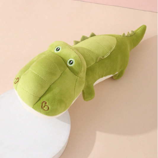 A stuffed crocodile toy sitting on a white plate. The crocodile is green and has a long tail. It is made of soft plush material and has a friendly expression.